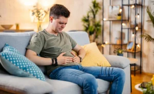 Man sitting on a couch at home holding his stomach in discomfort from bloating